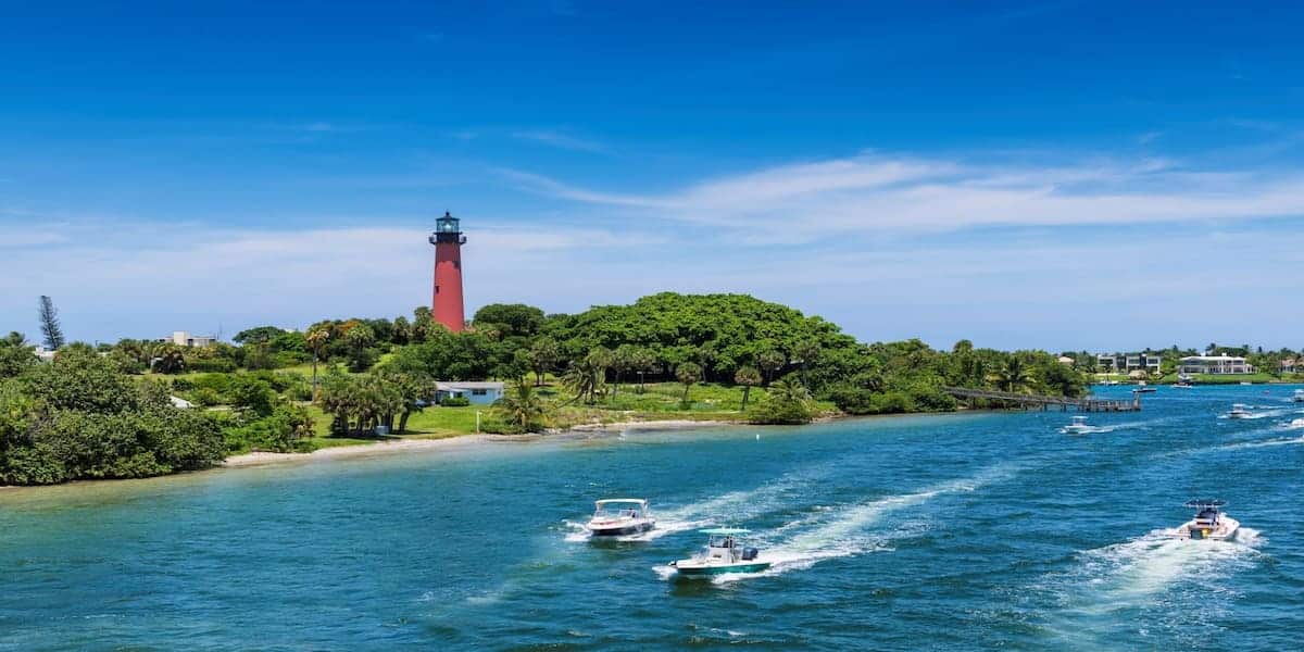 Boats cruising near a picturesque Florida lighthouse surrounded by greenery and blue waters, emphasizing the relevance of Florida life jacket laws for recreational boating safety.